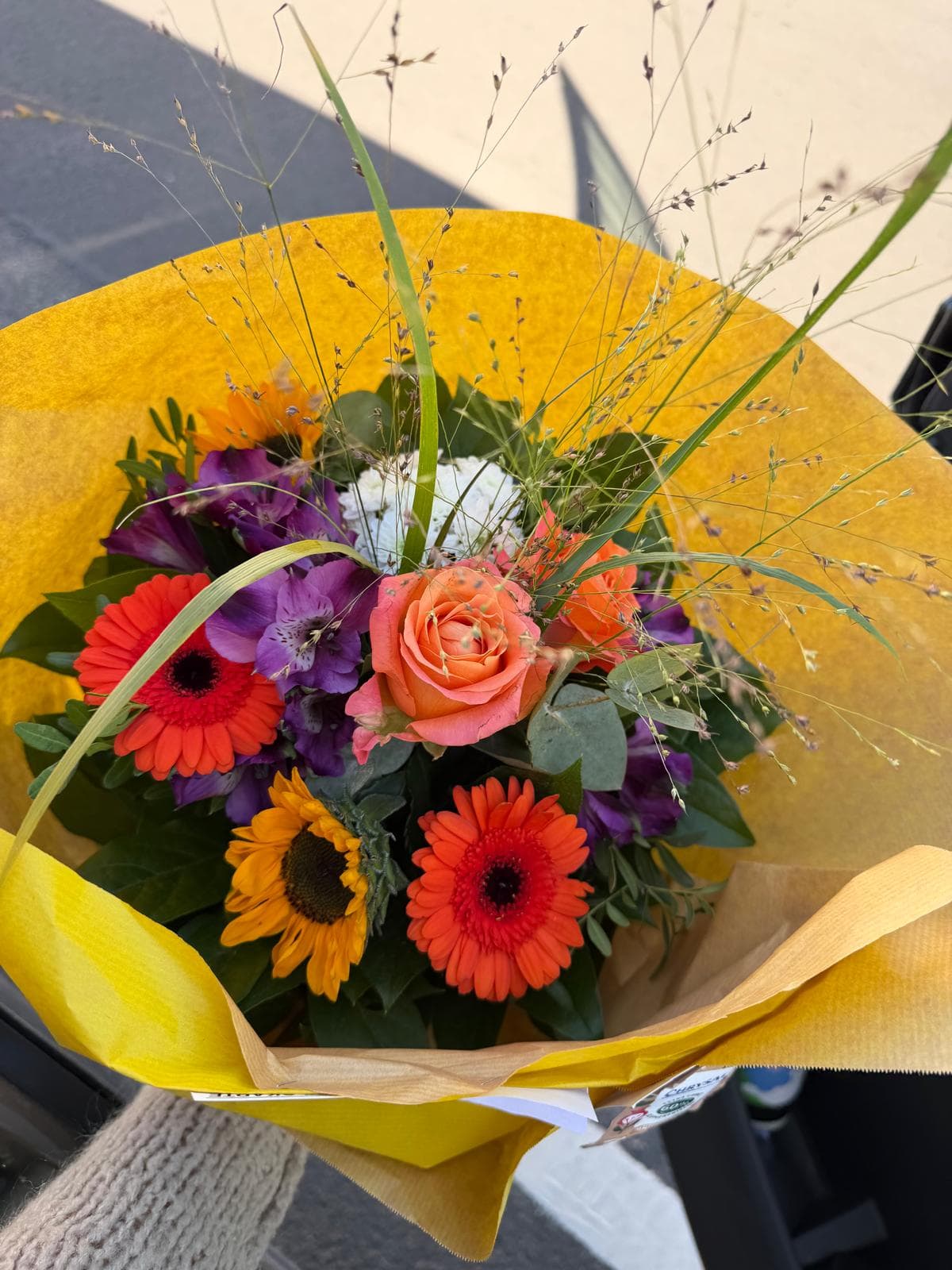 Bunter Blumenstrauß mit roten und orangefarbenen Gerberas, violetten Blüten, Rosen in Creme-Orange, Gräsern und Grünwerk, eingepackt in gelbes Papier