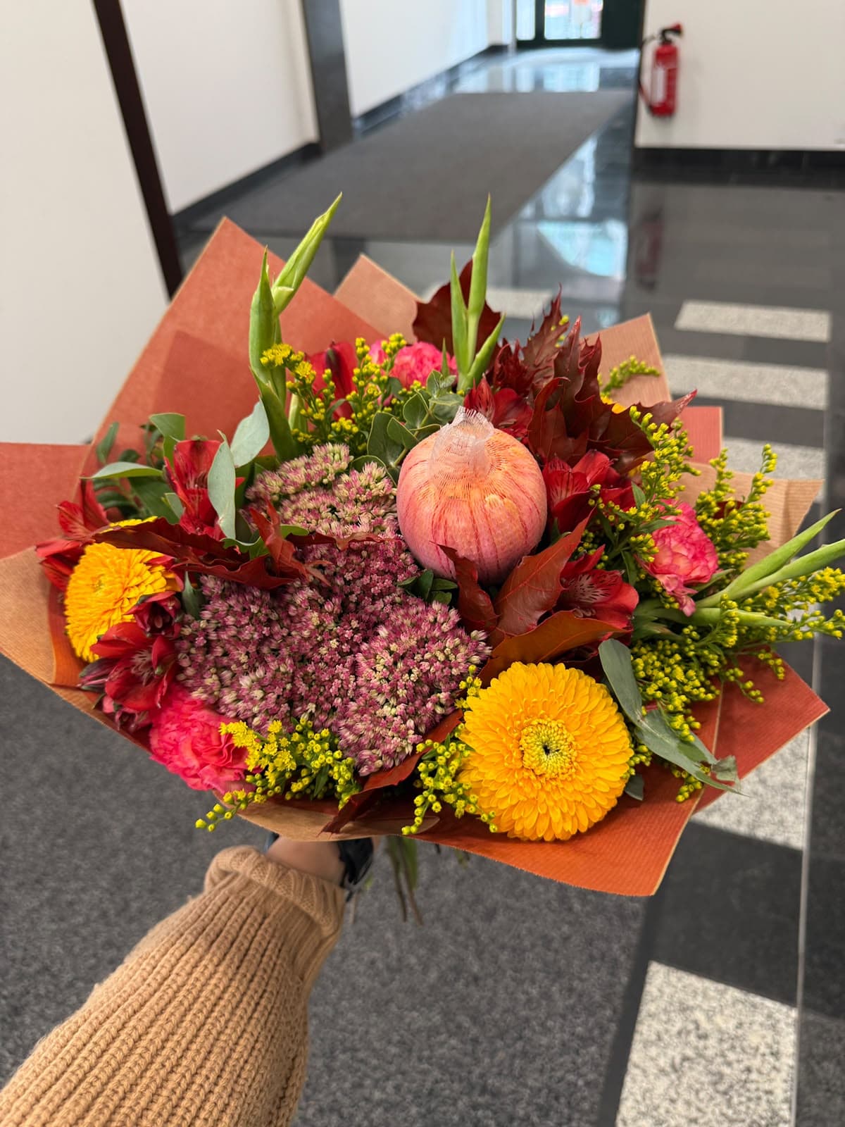 Bunter Blumenstrauß mit Gerbera, Nelke, Celosia und Trockenblu¨mchen in orange-roter Papierverpackung, von Hand gehalten.