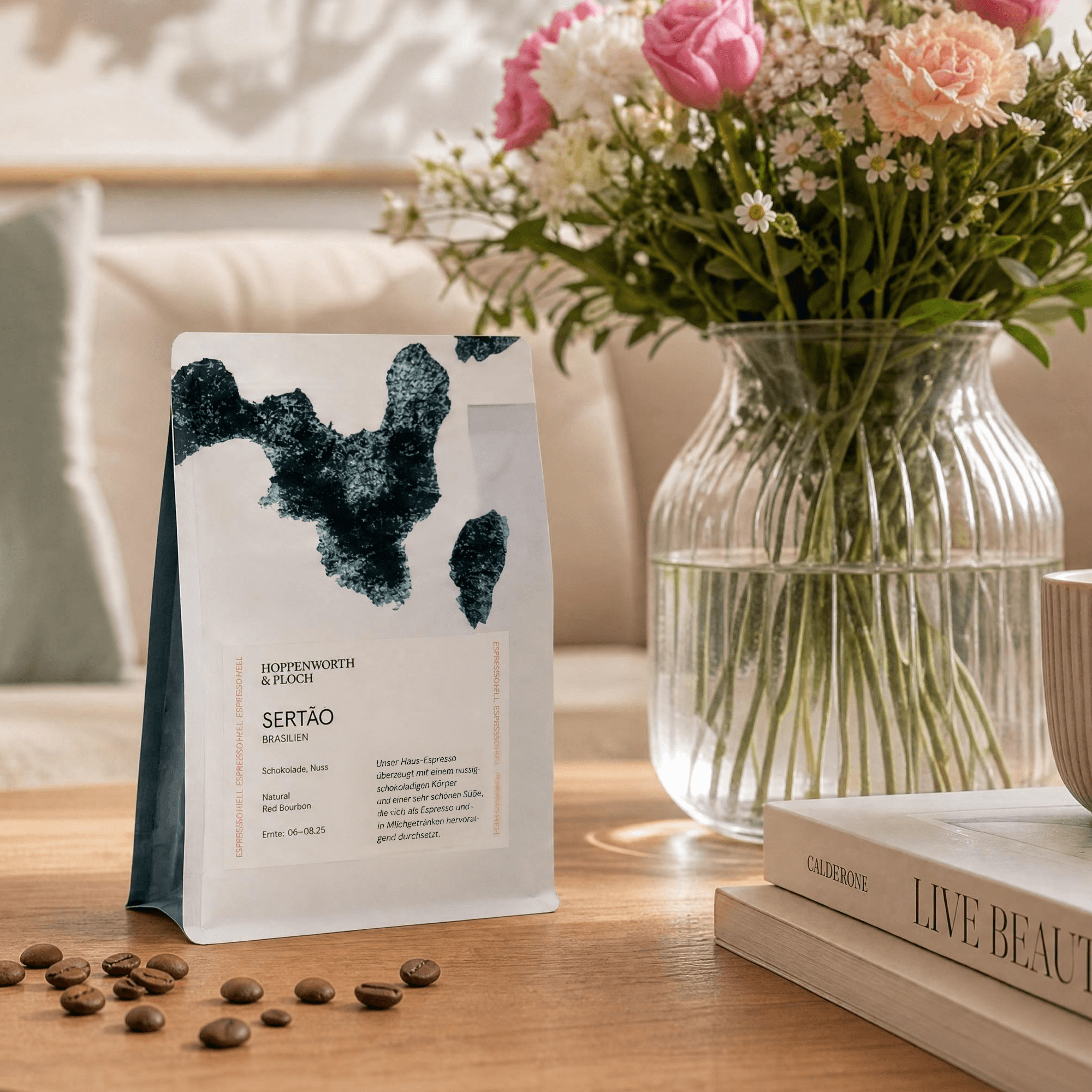 Weiße Kaffeetüte mit dunkelgrünem Muster neben klarer Vase mit pink-pfirsich farbenen Blüten und Büchern auf Holztisch